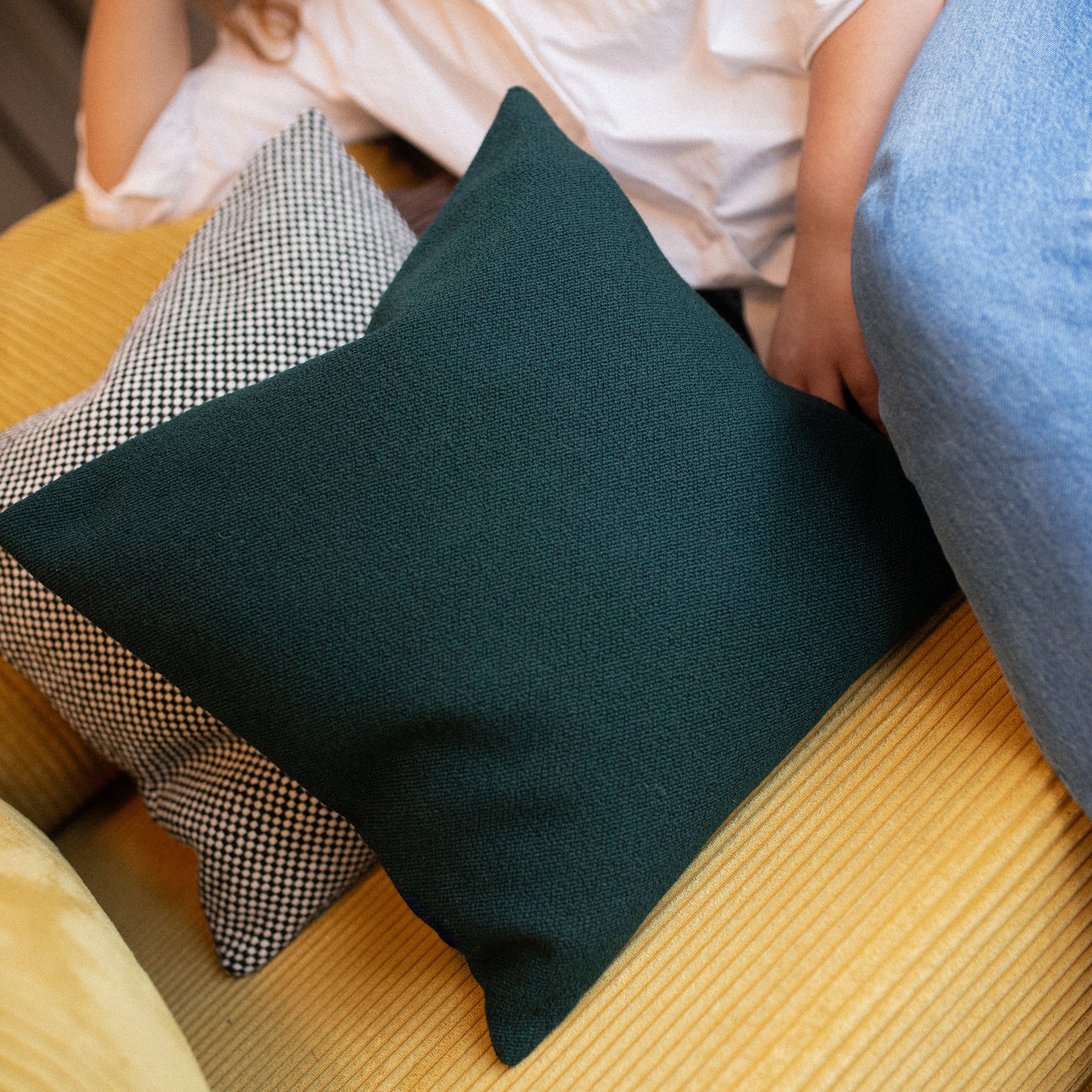 o puder i uld – en mørkegrøn og en sort-hvidternet – placeret på en sennepsgul sofa ved siden af en person iført jeans. Et varmt og taktilt interiørmotiv med fokus på tekstilernes struktur.