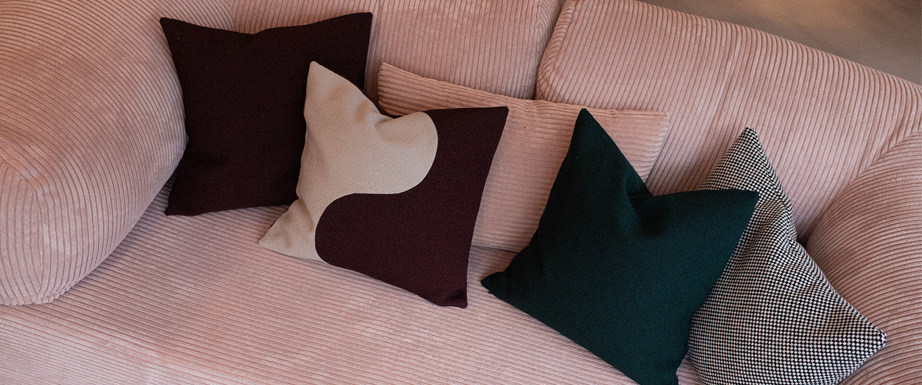 Omhu-sofa i støvet rosa fløjl med fire Marou-puder i bordeaux, beige, mørkegrøn og sort-hvidternet uld. Et harmonisk interiørbillede, der fremhæver kontrasten mellem farver og teksturer.